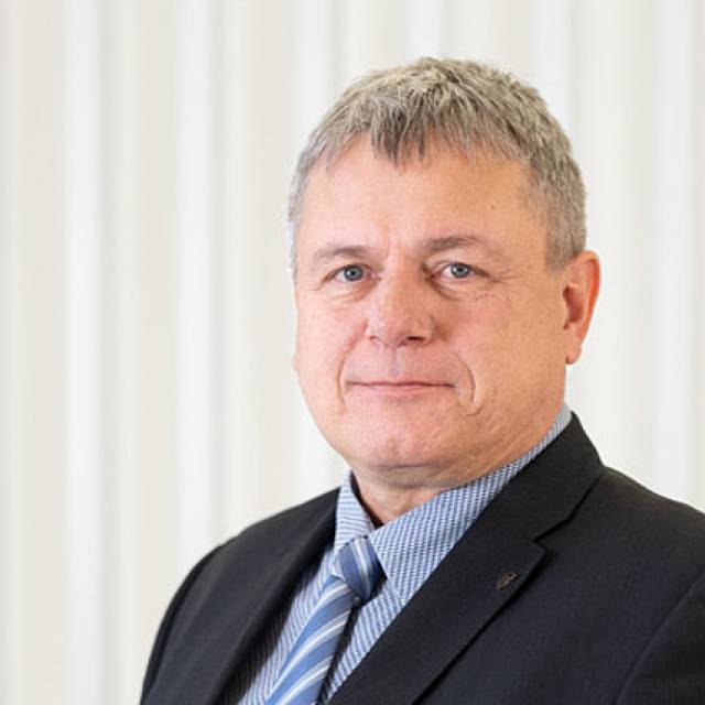 Portr&auml;t eines Mannes im Anzug mit Krawatte, grauem Haar, vor hellem Hintergrund.