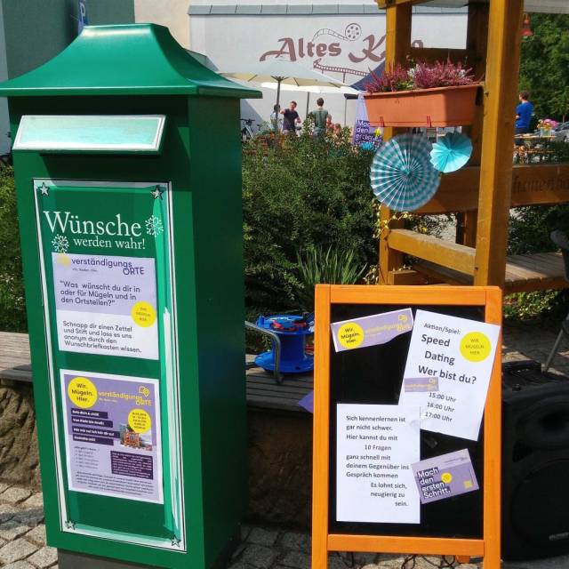 Grüner Wunschbriefkasten mit Schildern und Plakat „Speed Dating – Wer bist du?“.