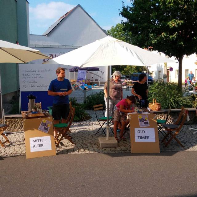 Menschen sitzen unter Sonnenschirmen an Tischen mit Schildern „Mittelalter“ und „Oldtimer“.